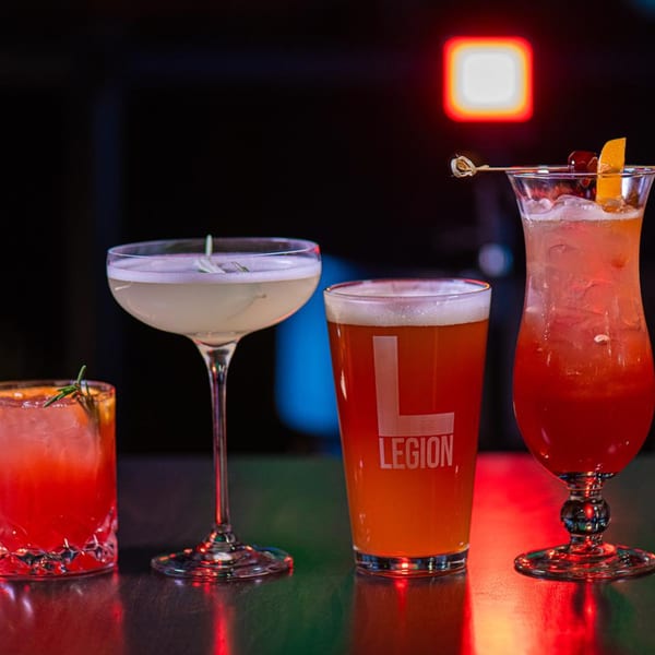 Four cocktails and a beer with the Legion logo on a bar top with colorful lighting.